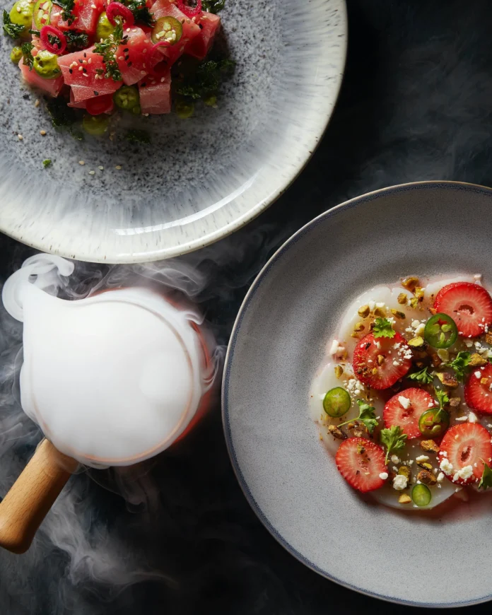 To the right side of the photo sits Scallop Carpaccio dressed in Chilled Strawberry Vinaigrette on a white plate.