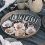 An array of 5 scallops, three baby octopus, and a bunch of sardines sit on a big round plate on a picnic table.