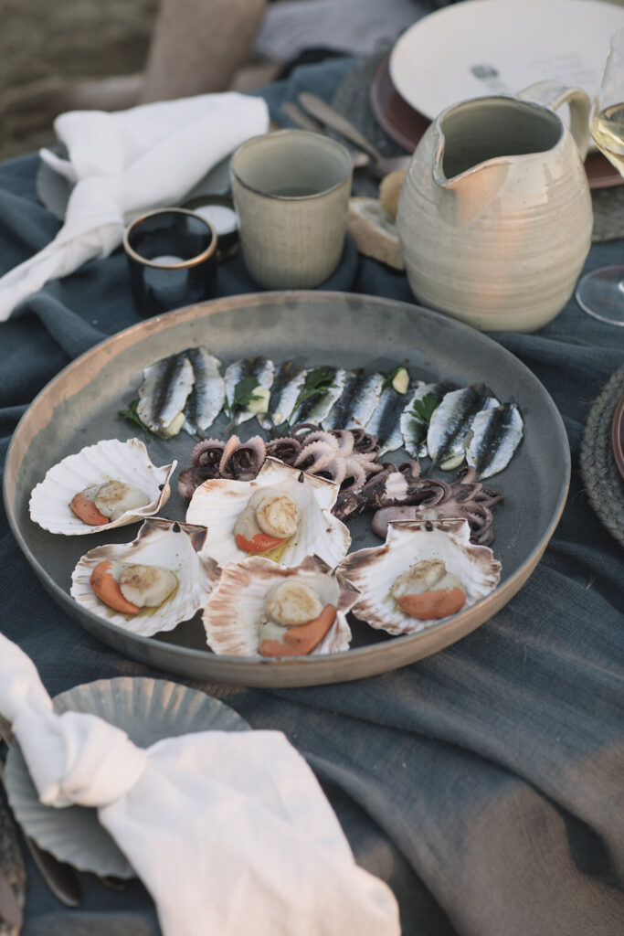An array of 5 scallops, three baby octopus, and a bunch of sardines sit on a big round plate on a picnic table.