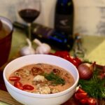 A white bowl of Seafood Chowder sits beside a glass of red wine, its bottle, garlic, onions, and tomatoes.