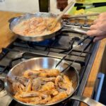 On a stovetop, a hand with tongs reaches into a steel pan filled with head-on shrimp to make 10 Minute Aegean-Style Shrimp with Caper Almond Pesto.