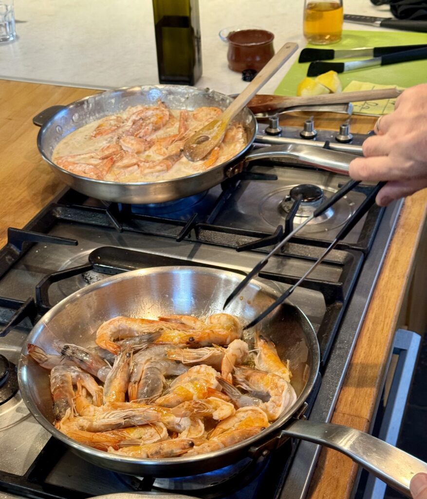 On a stovetop, a hand with tongs reaches into a steel pan filled with head-on shrimp to make 10 Minute Aegean-Style Shrimp with Caper Almond Pesto.
