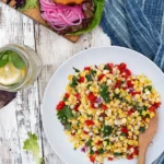 Corn, peppers, and onions mixed together in a Sweet Corn Relish on a white plate with a brown wooden spoon in the mixture.