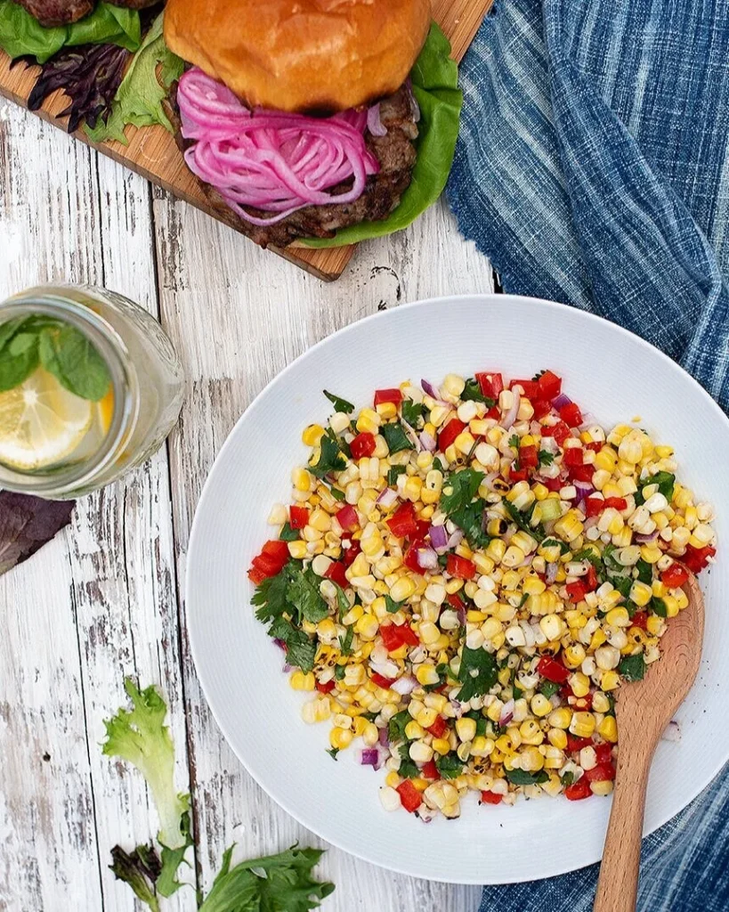 Corn, peppers, and onions mixed together in a Sweet Corn Relish on a white plate with a brown wooden spoon in the mixture.