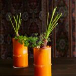 In two highball glasses sits Carrot Bloody Mary cocktails, orange in color with celery sticks as garnish