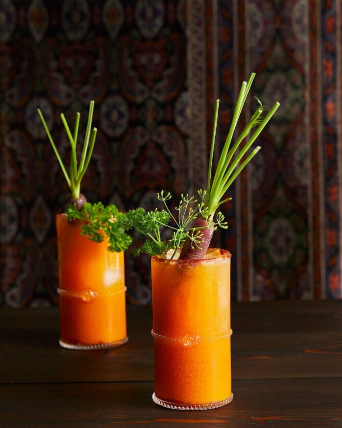 In two highball glasses sits Carrot Bloody Mary cocktails, orange in color with celery sticks as garnish