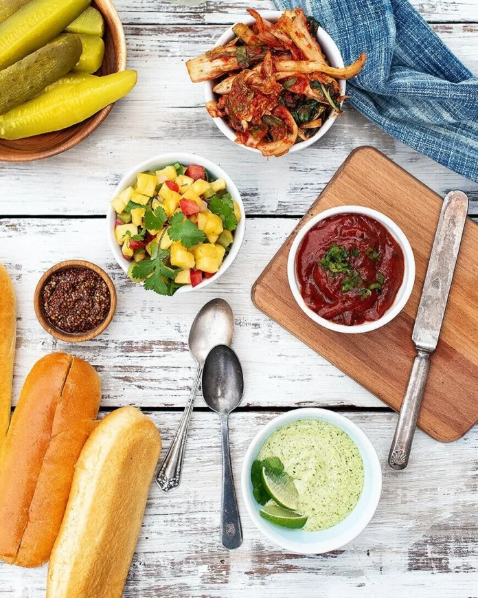 Three small bowls of condiments sit on a white picnic table with spoons and hot dog buns spread across the table too.