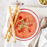 An above shot of Tomato Strawberry Gazpacho, pink-red in color, served in a bowl with three thin breadsticks on the side.