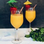 Two tall glasses with stems hold Tropical Mango Smoothie with strawberries on the rim with little cocktail umbrellas in the strawberries.