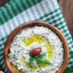 A brown bowl of tzatziki sits on a green and white kitchen towel.