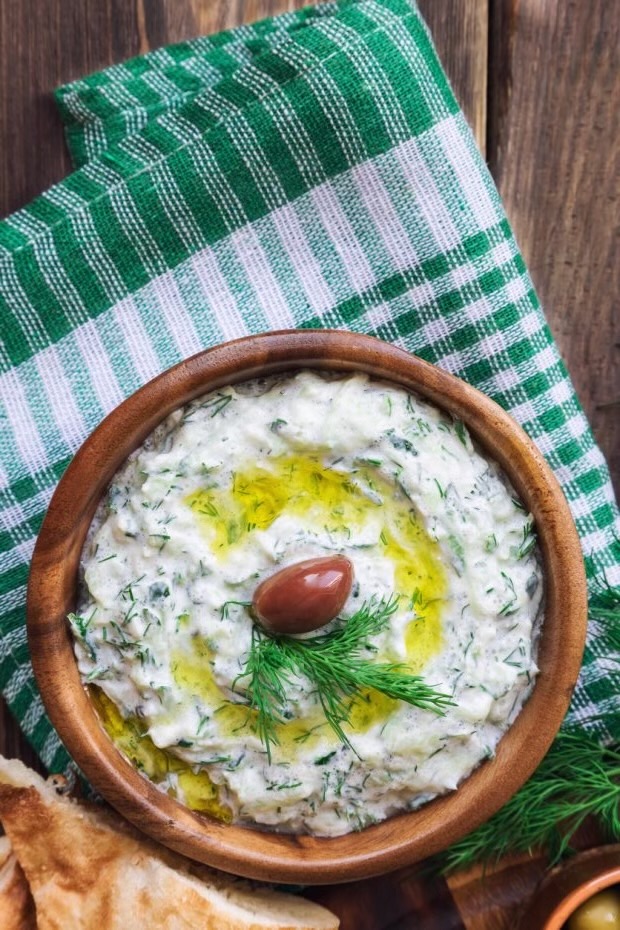 A brown bowl of tzatziki sits on a green and white kitchen towel.