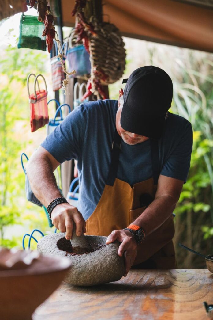 A person in a black cap and brown apron grinds spices in a stone mortar and pestle on a wooden surface.
