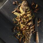 On a black wooden surface is a Rainbow Trout with Fennel and Mushrooms sitting next to a knife.