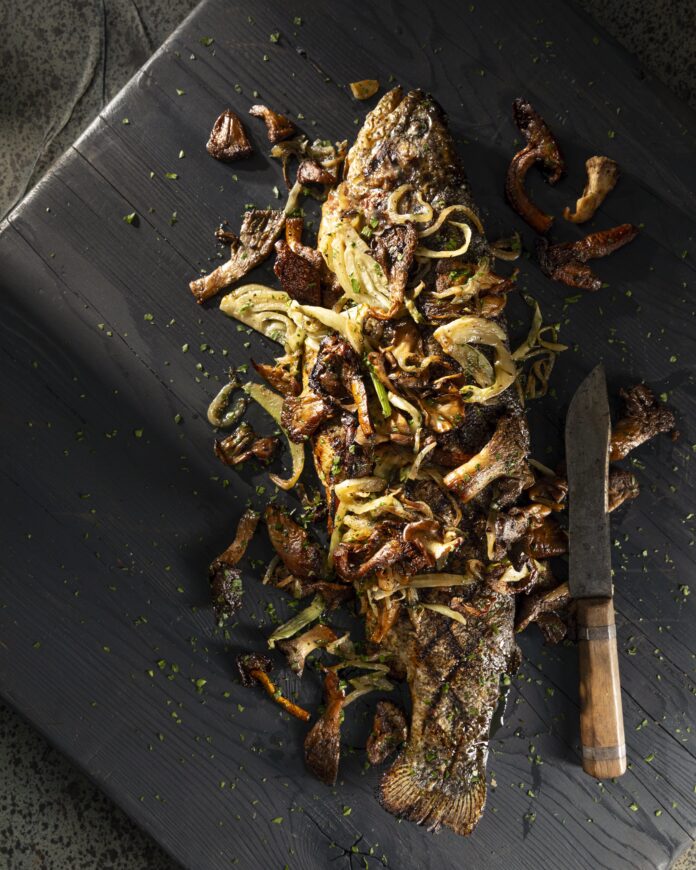 On a black wooden surface is a Rainbow Trout with Fennel and Mushrooms sitting next to a knife.