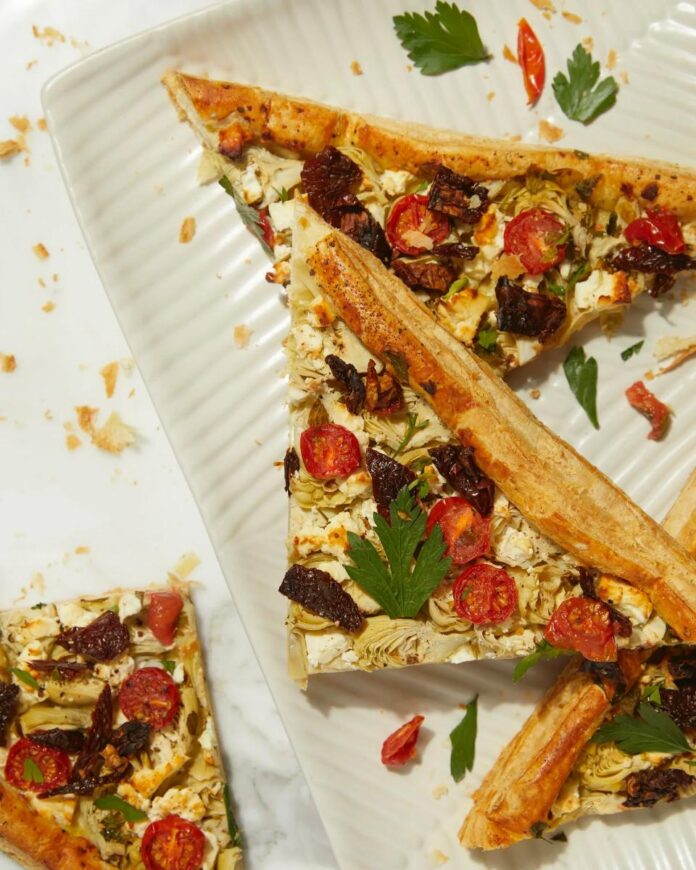 A flat lay photo of several slices of a savory artichoke puff pastry tart, topped with sun-dried tomatoes, feta cheese, and parsley.
