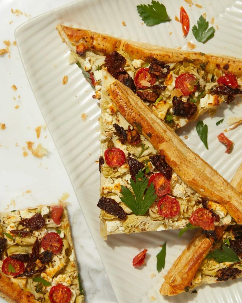 A flat lay photo of several slices of a savory artichoke puff pastry tart, topped with sun-dried tomatoes, feta cheese, and parsley.