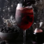 A wine glass holds a Blackberry Crush Spritz with blackberries in the liquid and as a garnish all against a dark background.