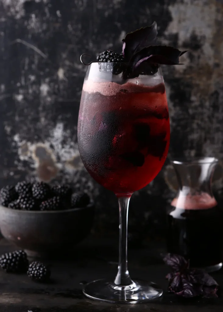 A wine glass holds a Blackberry Crush Spritz with blackberries in the liquid and as a garnish all against a dark background.