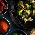 A plate of roasted broccoli florets with brown butter bread crumbs, surrounded by bowls of salt, red pepper flakes, paprika, and more broccoli.
