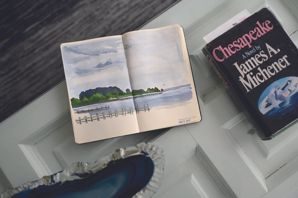 A painting of the Chesapeake Bay in a sketchbook on a table beside another book.