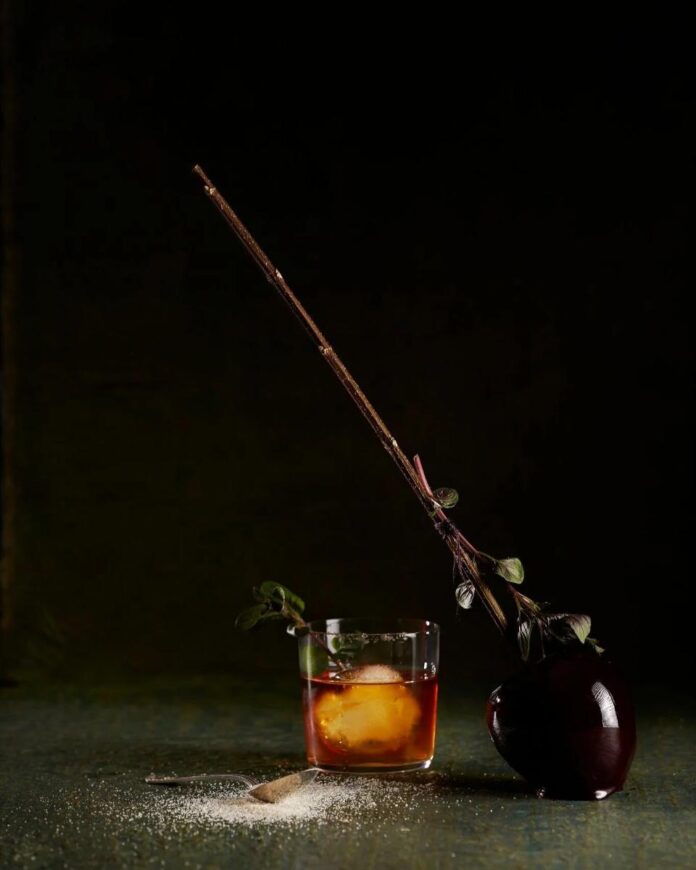 A Coffee Old Fashioned cocktail in a rocks glass with a large ice cube, garnished with a leafy branch, next to a caramel apple on a stick.