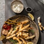 Deep fried dilly beans on a brown plate with a side of garlic aioli sitting in a small brown dish beside it.