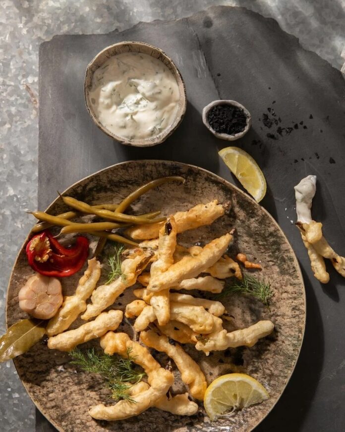 Deep fried dilly beans on a brown plate with a side of garlic aioli sitting in a small brown dish beside it.