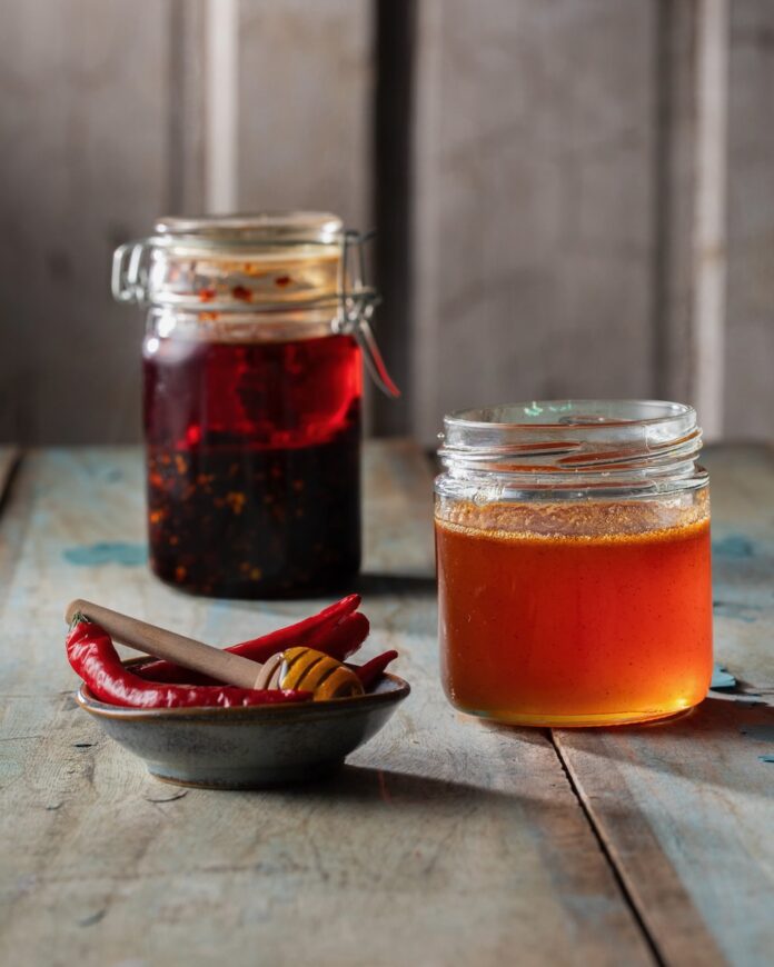 A jar of easy homemade hot honey sits beside a jar of chili crisp and a small bowl of red chiles.