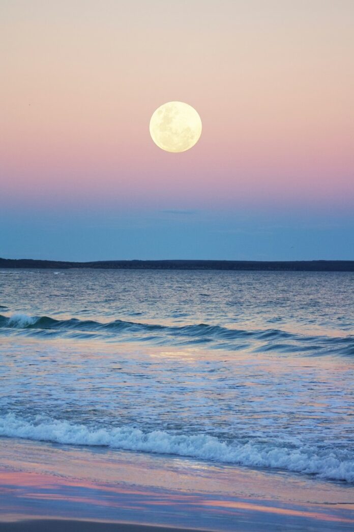 A full moon sits above the ocean rolling into the August sands as the sun sets.