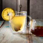 A clear mug with a Hot Honey Hot Toddy inside featuring a metal spoon, cinnamon stick, and lemon round.