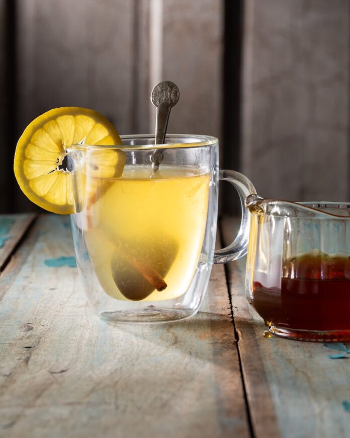 A clear mug with a Hot Honey Hot Toddy inside featuring a metal spoon, cinnamon stick, and lemon round.