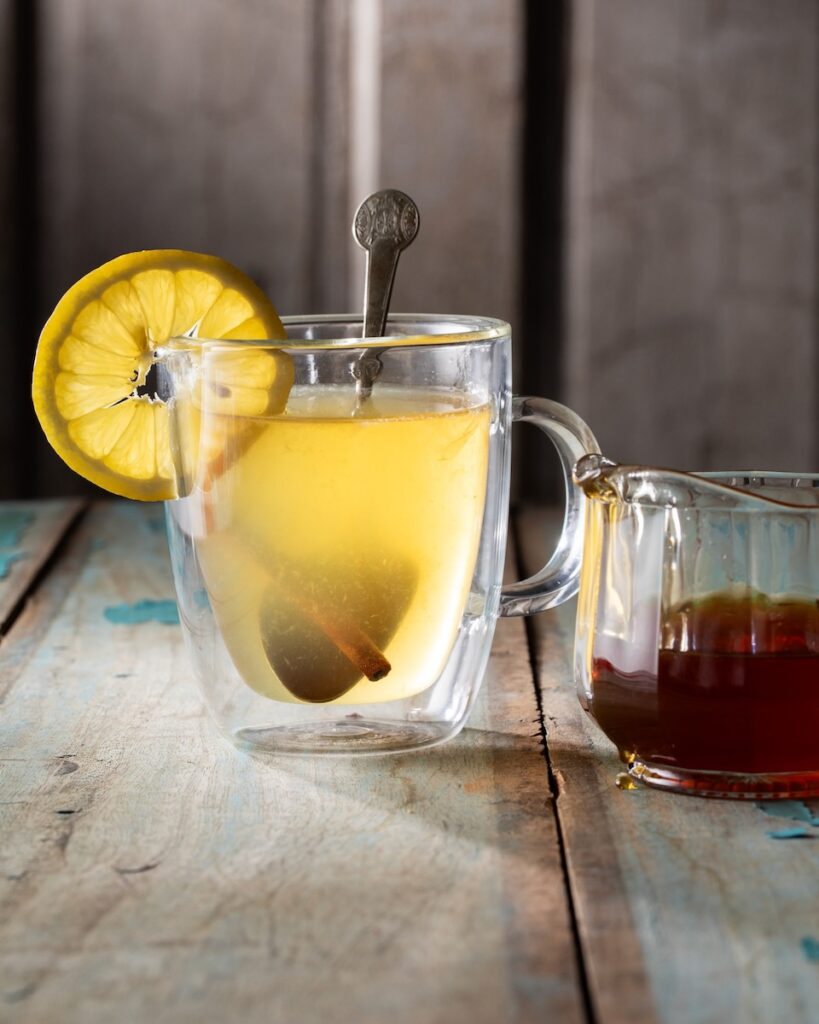 A clear mug with a Hot Honey Hot Toddy inside featuring a metal spoon, cinnamon stick, and lemon round.
