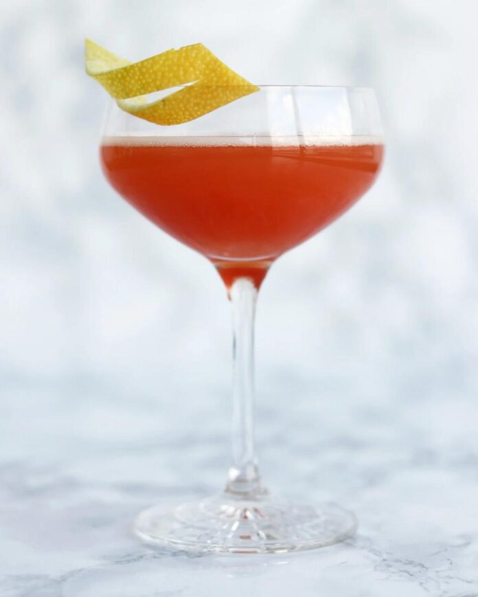 A stemmed glass filled with a pinkish-red Jack Rose cocktail, garnished with a spiral of lemon peel.