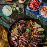 A chicken fajita skillet with slices chicken, limes, and peppers. Then there are small bowls of pico, sour cream, and onions.