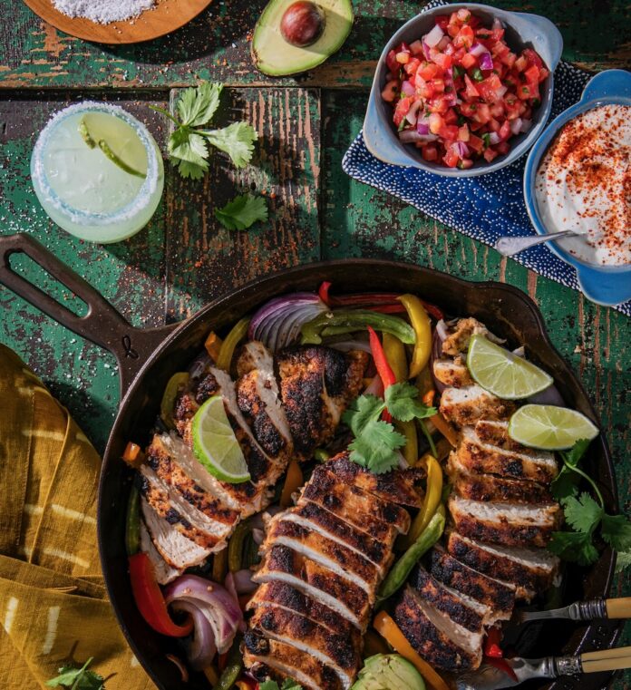 A chicken fajita skillet with slices chicken, limes, and peppers. Then there are small bowls of pico, sour cream, and onions.