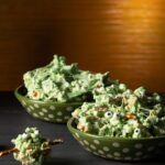 Two green bowls with white polka filled with green marshmallow covered popcorn with pretzel sticks and candy eyes.