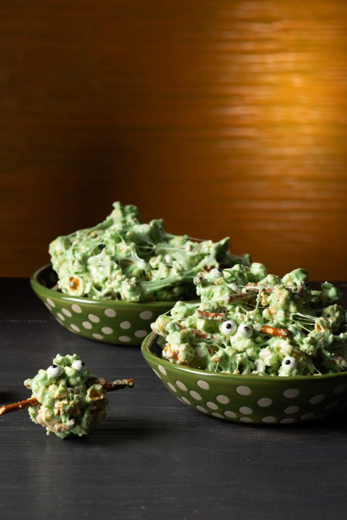 Two green bowls with white polka filled with green marshmallow covered popcorn with pretzel sticks and candy eyes.