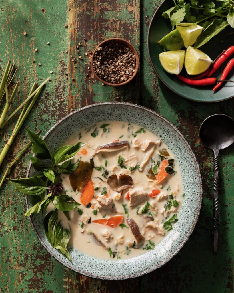 A grey bowl full of a Martha Stewart Slow Cooker Tom Kha Gai with leafy greens garnish sits on a green picnic table.