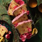 A sliced into pieces whole lemon pound cake sits with a pomegranate glaze on top with pink flowers and pomegranate seeds as decor.