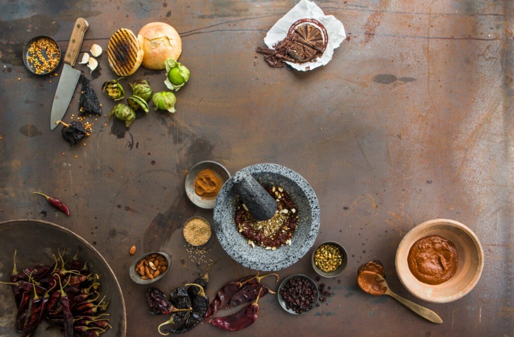 A flat lay photo showing ingredients for Mexican mole sauce, including various chiles, nuts, spices, and a stone mortar and pestle on a rustic metal surface.