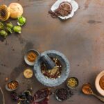 A flat lay photo showing ingredients for Mexican mole sauce, including various chiles, nuts, spices, and a stone mortar and pestle on a rustic metal surface.