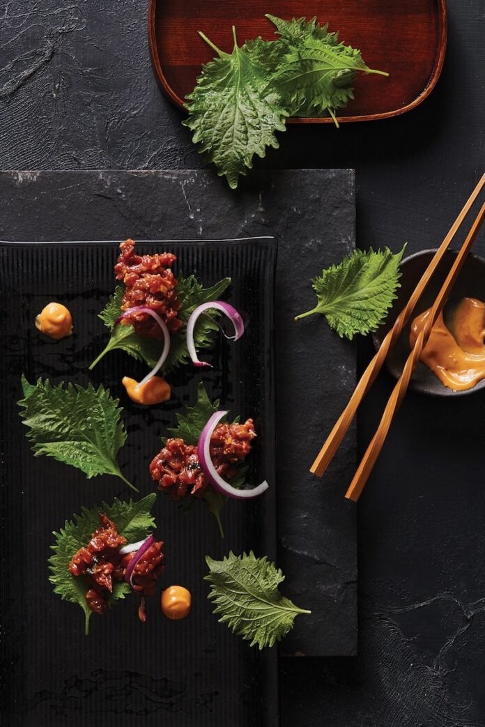 Various bites of raw dry-aged beef Ssam with leafy greens and small pieces of beef as chopsticks sit off to the side.