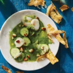 A smooth green aguachile with scallops and chips in a white bowl on a blue table.
