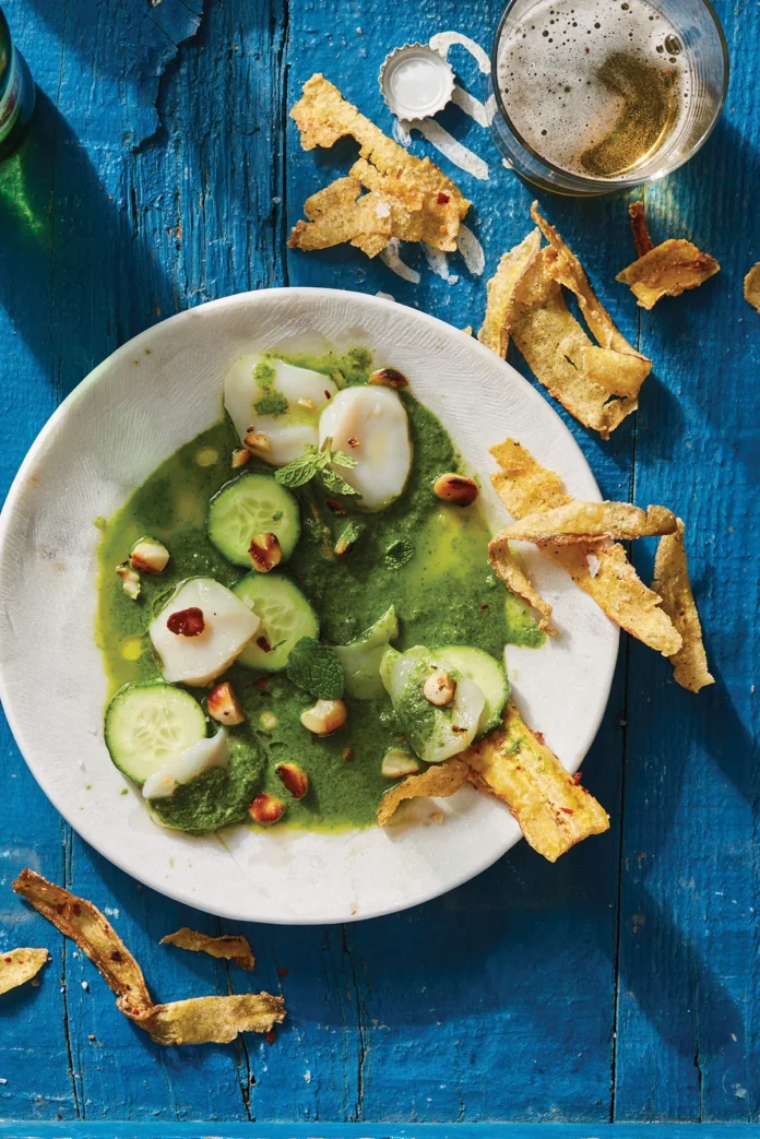 A smooth green aguachile with scallops and chips in a white bowl on a blue table.