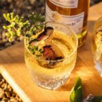 A Smoked Maple and Scotch Old Fashioned cocktail in a rocks glass, garnished with grilled peaches and fresh herbs, with a bottle of Scotch in the background.
