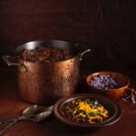 Texas chili sits in a large pot with a bowl of chili in front of it topped with cheese and a bowl of red onions beside it.