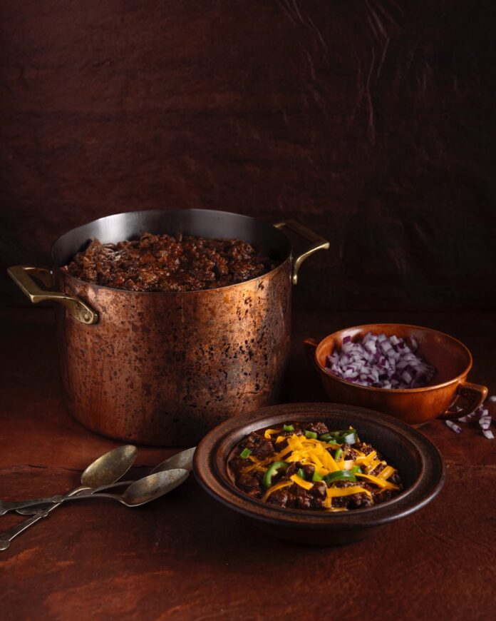Texas chili sits in a large pot with a bowl of chili in front of it topped with cheese and a bowl of red onions beside it.