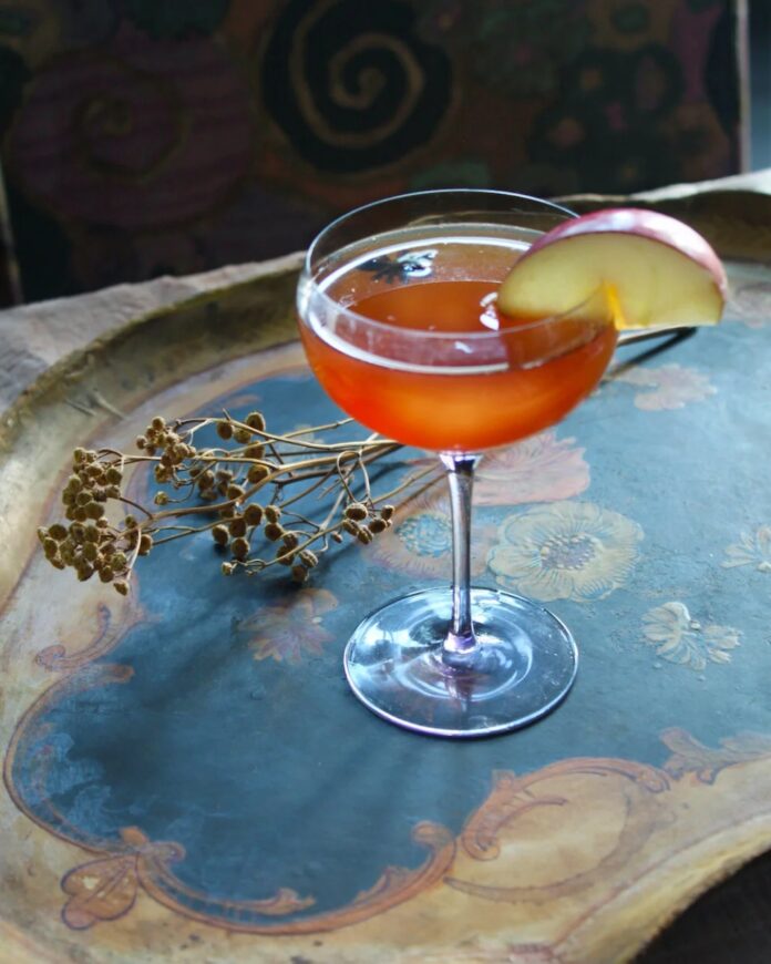A reddish-orange cocktail in a coupe glass, garnished with a thin apple slice, and resting on a rustic serving tray with dried foliage.
