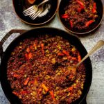 A cast iron skillet with toasted red quinoa and pickled vegetables, with two black bowls and a fork on the side.