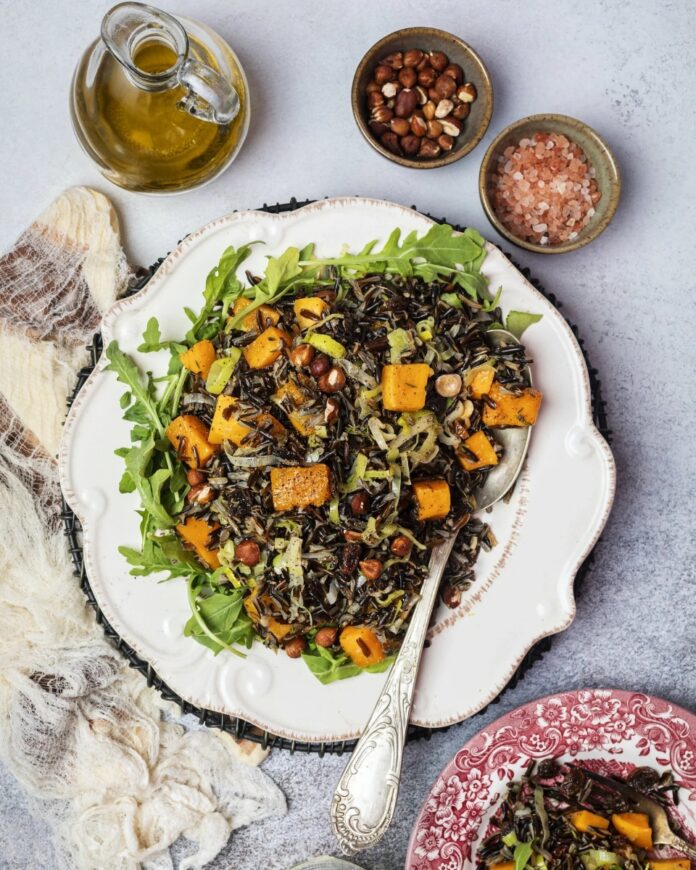 A plate of wild rice with butternut squash, raisins, and hazelnuts, served over a bed of arugula, next to a bottle of olive oil and bowls of salt and hazelnuts.